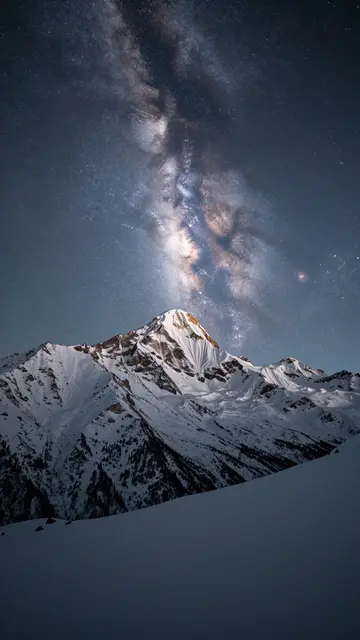 Snow-capped mountain peaks stand beneath a bright glowing milky way in the dark night sky