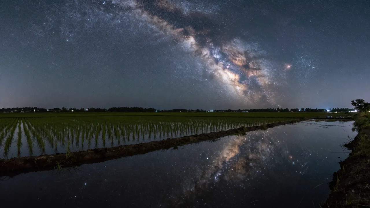 The glowing milky way stretches across the night sky, mirrored in a flooded rice field