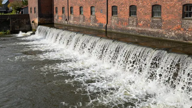 White water spills over a wide river weir alongside an aged red brick mill building