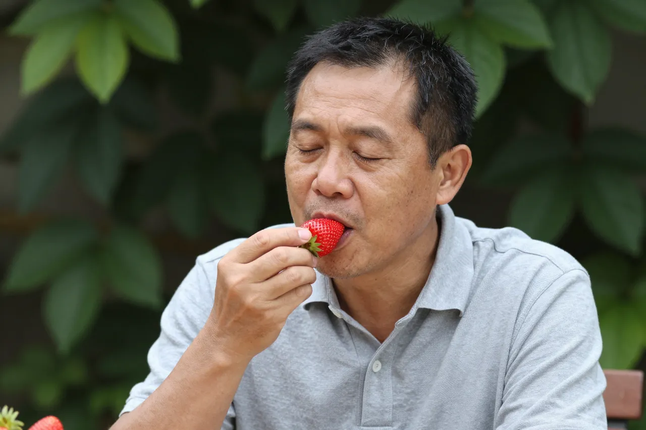 Middle-aged Southeast Asian man with closed eyes eats a fresh strawberry outdoors in a garden