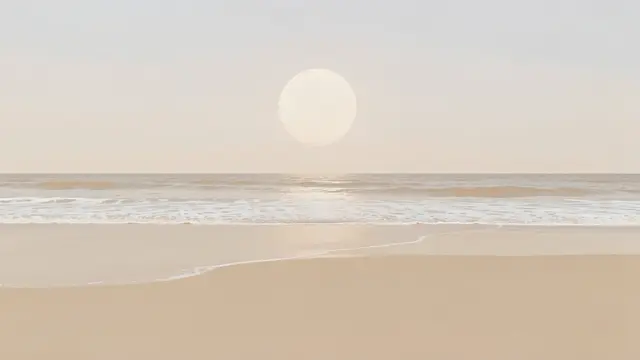 Pale soft sun hangs over a quiet beige beach with thin gentle surf along the shore