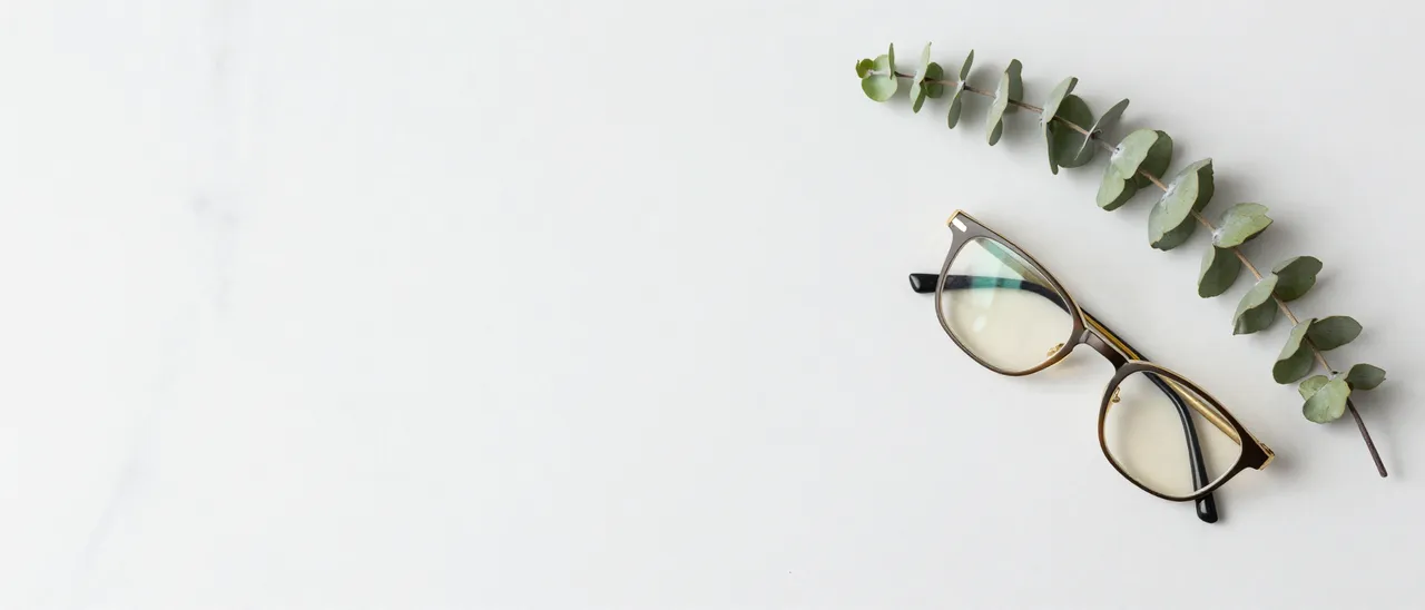 Top view of a white marble desk surface with reading glasses and a eucalyptus sprig in the right corner, with wide empty
