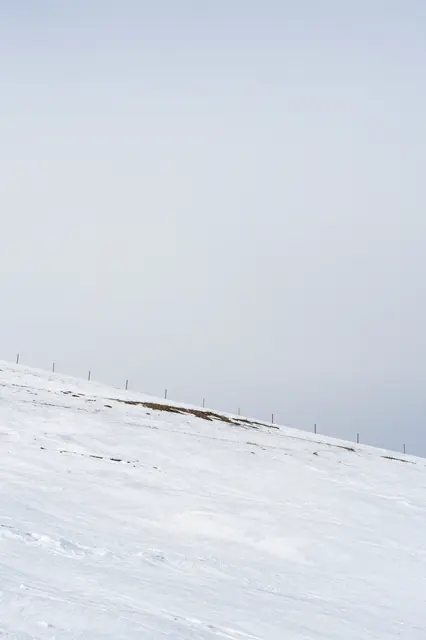 A steep snowy hillside with a line of fence posts under a vast pale overcast sky and empty upper copy space.