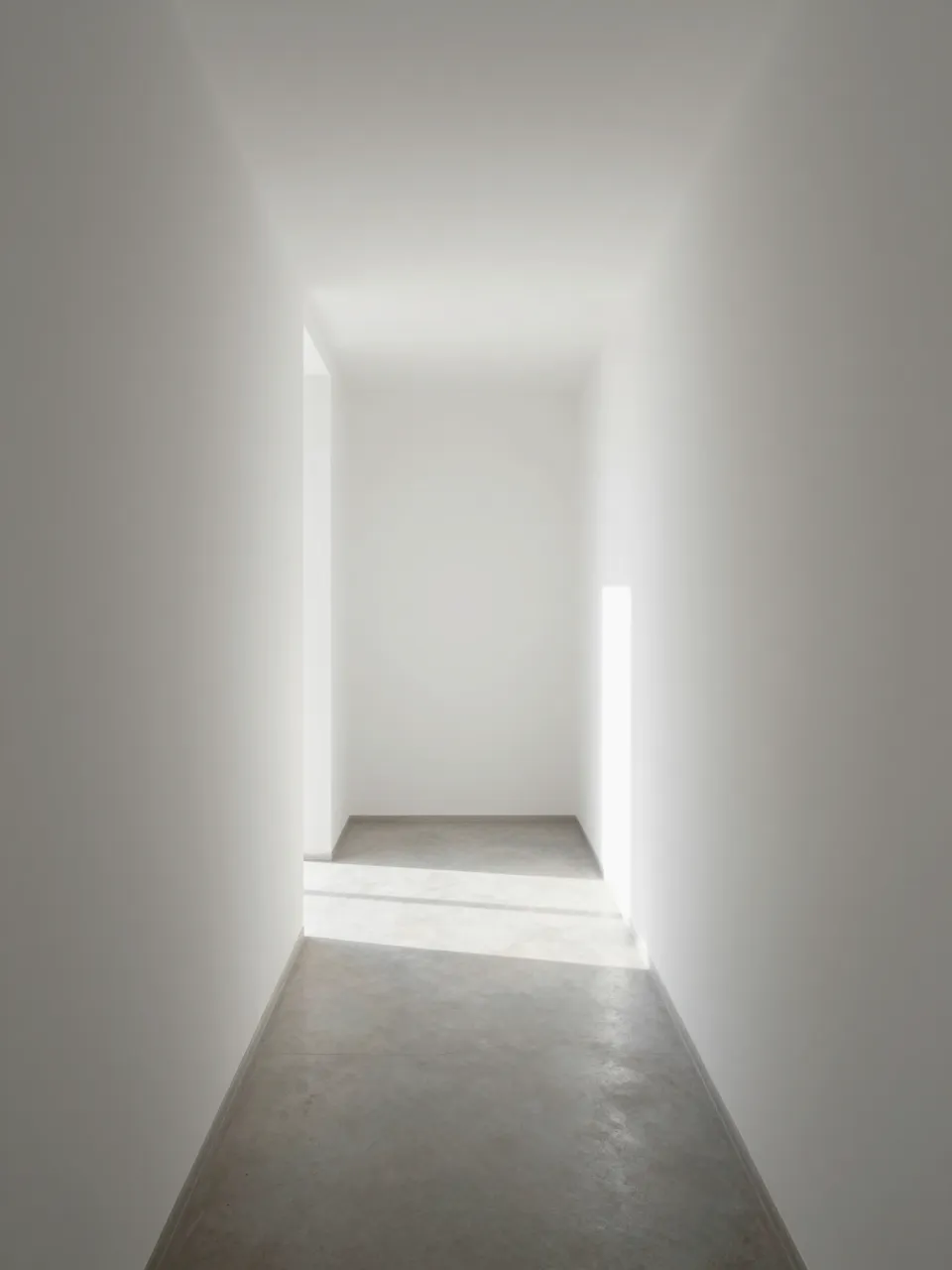 Narrow sunlit minimalist white corridor with concrete floor and soft light at the far end