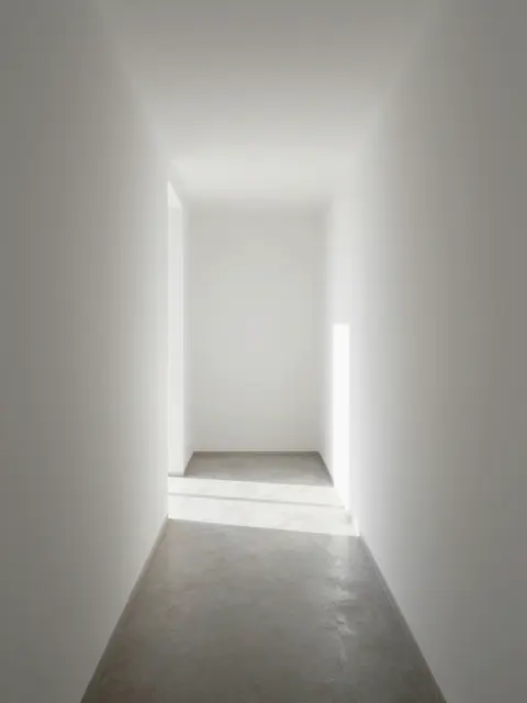 Narrow sunlit minimalist white corridor with concrete floor and soft light at the far end