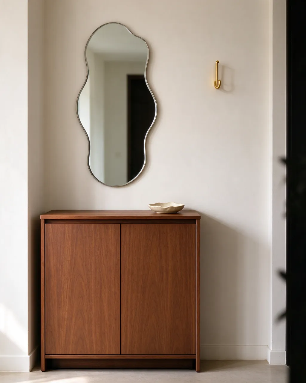 A narrow wooden entry cabinet sits below a wavy wall mirror beside a brass hook and small ceramic dish on a neutral wall.