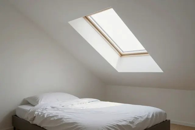 A low bed with white linens sits beneath a wooden frame skylight in a bright sloped-ceiling attic bedroom.