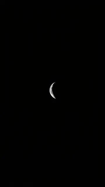 A thin pale crescent moon sits centered against a solid deep black night sky