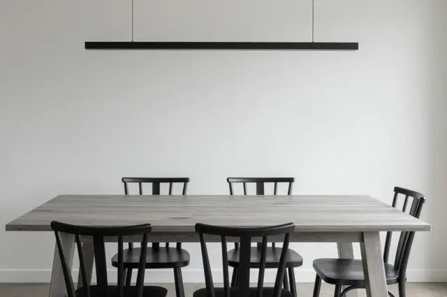 A long light ash dining table with black spindle chairs sits below a linear black pendant light against a plain white wall