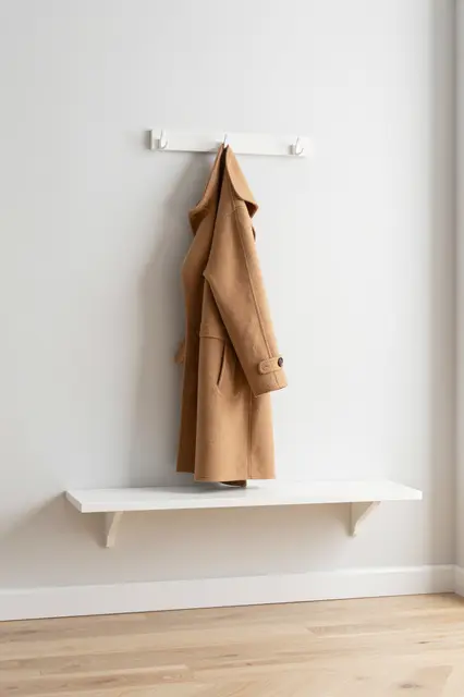 A camel wool coat hangs from a wall hook above a floating white shelf next to pale oak flooring