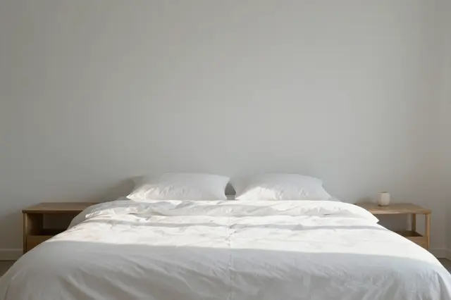 A minimalist bedroom with a rumpled white bed lit by soft morning sunlight