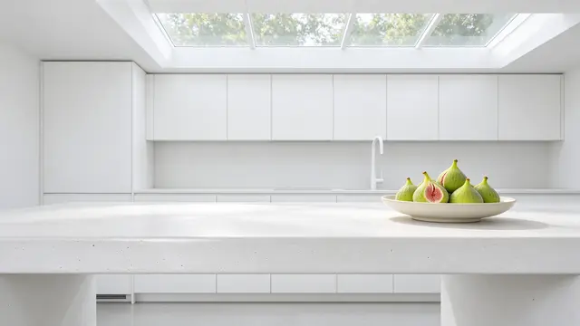 An all-white minimalist kitchen displays a bowl of fresh green figs on a microcement island under a long overhead skylight.