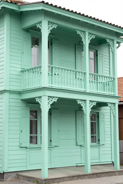 Corner view of a two-story narrow mint green wooden house with ornate porch brackets and shuttered windows