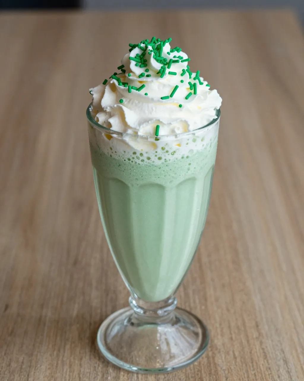 Tall glass of mint milkshake topped with whipped cream and green sprinkles sits on a wooden table