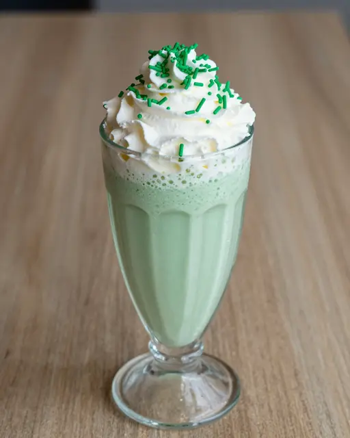 Tall glass of mint milkshake topped with whipped cream and green sprinkles sits on a wooden table