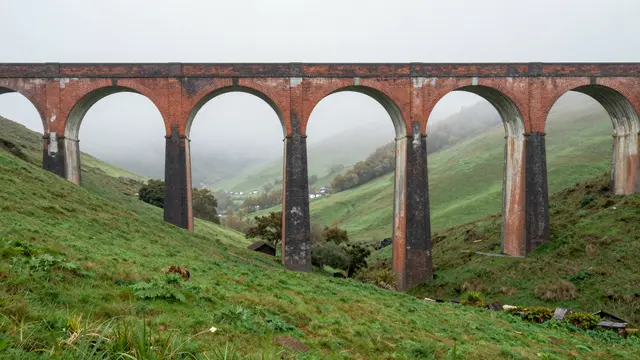 Red brick arched railway viaduct spans a lush green valley under foggy overcast morning skies