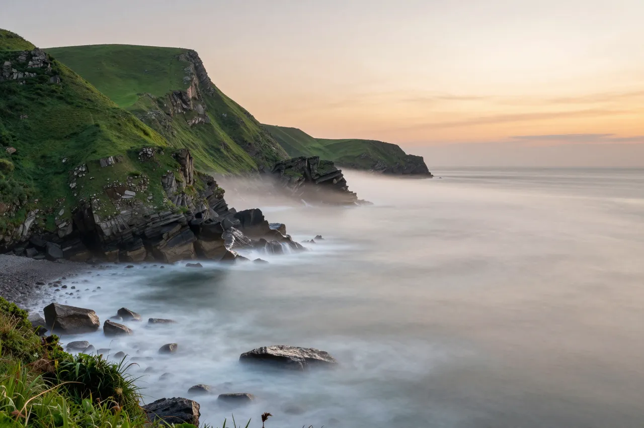Long exposure of sea fog drifting over green coastal cliffs at warm sunset