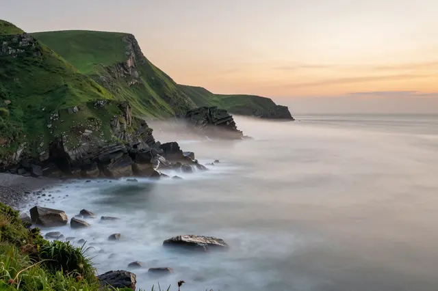 Long exposure of sea fog drifting over green coastal cliffs at warm sunset