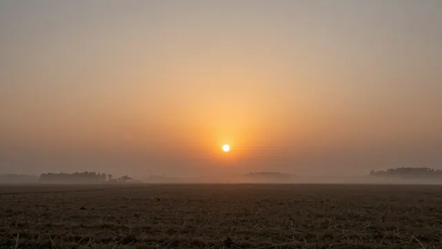 Glowing orange sun rises over fog-shrouded harvested farm fields at dawn