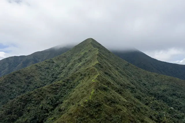 A steep grassy green mountain peak rises with low clouds wrapping surrounding summits under an overcast sky
