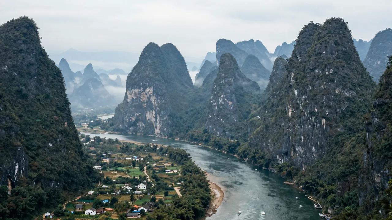 A high-angle view of steep forested karst peaks rising above a winding misty river valley with a small village.