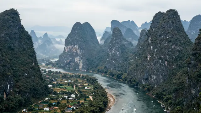 A high-angle view of steep forested karst peaks rising above a winding misty river valley with a small village.