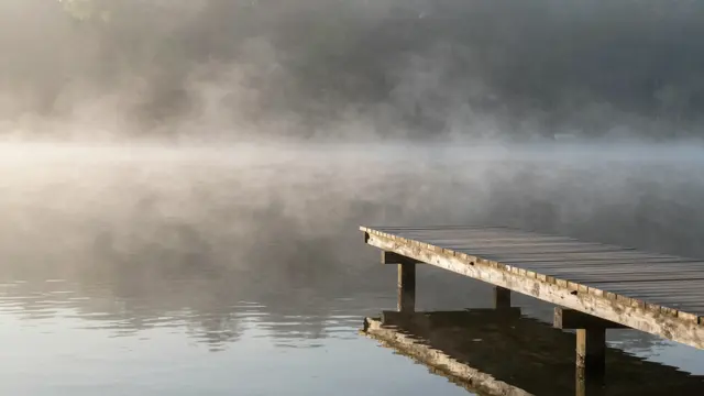 A weathered wooden jetty extends into a calm misty lake at soft golden morning light