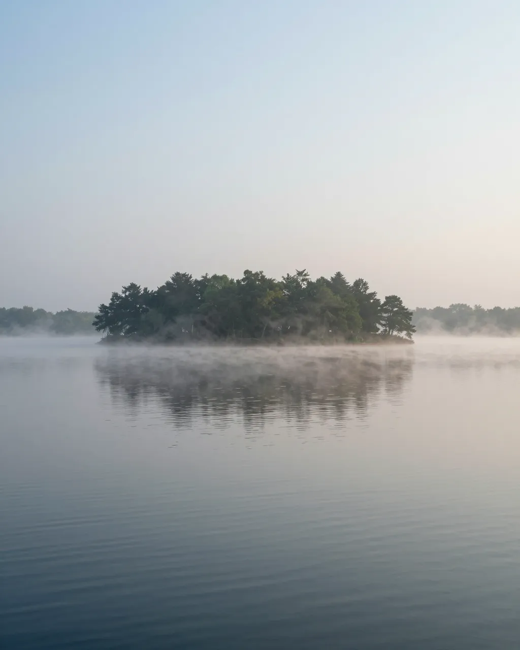 Calm misty lake with a forested island and wide empty sky above still water
