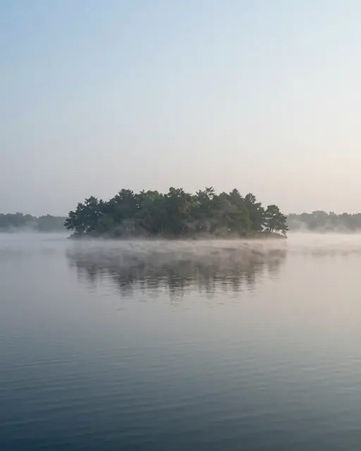 Calm misty lake with a forested island and wide empty sky above still water
