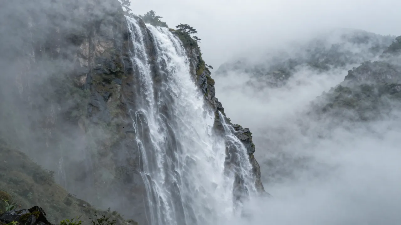 A tall cascading waterfall pours over a rocky cliff shrouded in thick rolling mountain fog