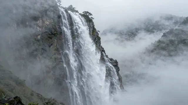 A tall cascading waterfall pours over a rocky cliff shrouded in thick rolling mountain fog
