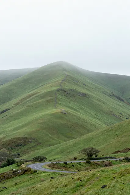 Winding country road cuts through grassy mist-covered hills under an overcast foggy sky