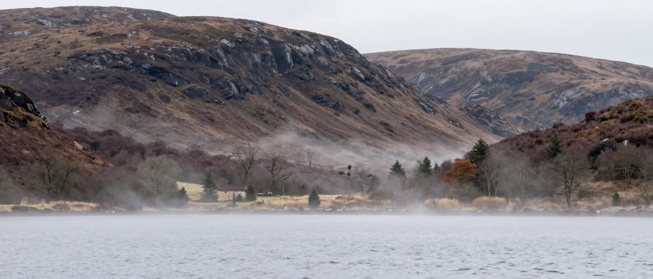 Fog drifts over a calm loch below rocky heather-covered highland hills under an overcast sky