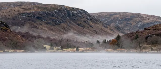Fog drifts over a calm loch below rocky heather-covered highland hills under an overcast sky