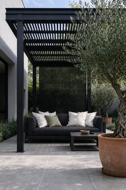 Black steel pergola shades an outdoor sofa on a stone terrace with potted olive trees