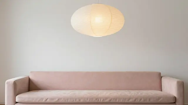 A soft blush velvet sofa sits below a glowing round pleated paper pendant light against a plain pale wall.