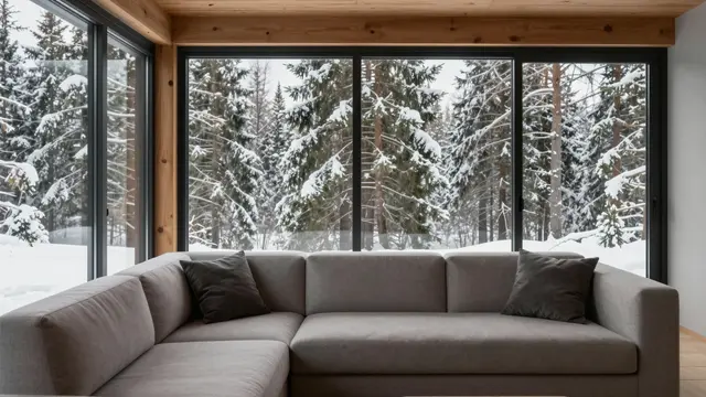 Gray sectional sofa sits in a wooden cabin interior overlooking snow-covered pine trees through large glass windows
