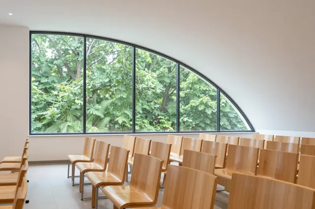 Rows of light wood chairs sit beneath a curved arched window showing green leafy trees outside