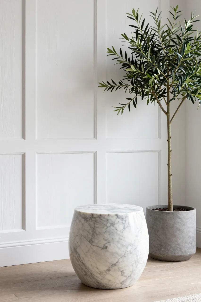 Marble drum side table and potted tall olive tree sit beside white paneled wall in a bright modern room