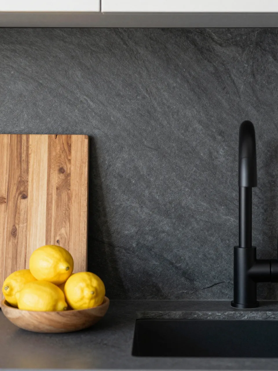 Dark slate kitchen backsplash with black faucet, wooden cutting board and a bowl of fresh lemons on the countertop