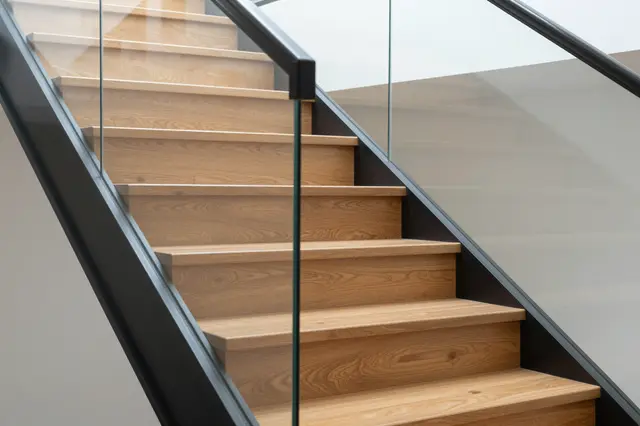 A partial view of a modern interior staircase with light wood steps and a glass and metal handrail