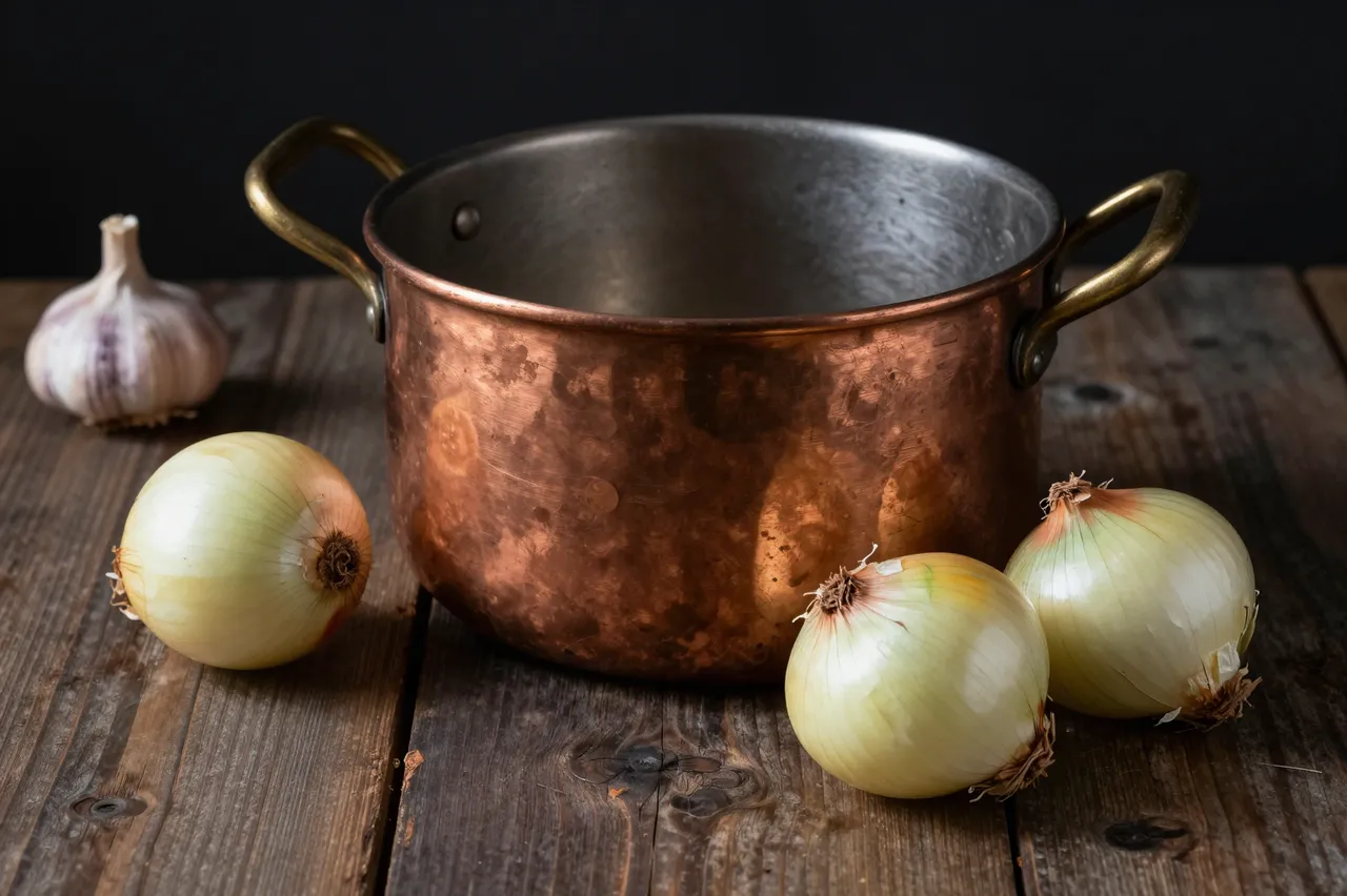Worn copper cooking pot sits with yellow onions and a garlic head on a dark rough wooden table