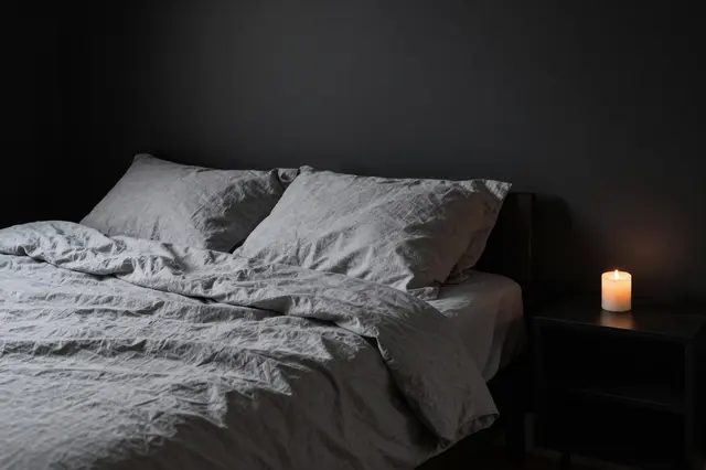 A dim moody bedroom with rumpled gray linen bedding and a lit candle on a black nightstand against dark walls