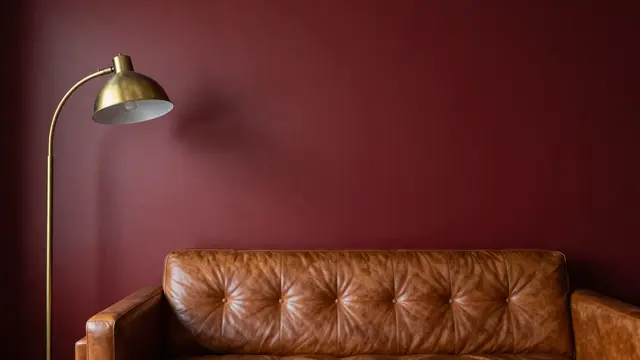 A tufted brown leather sofa and brass floor lamp sit against an oxblood red wall with large empty copy space.