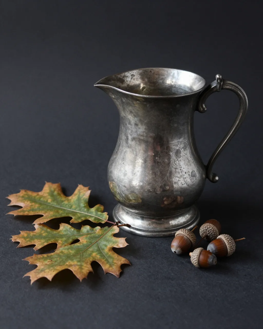 An aged engraved pewter jug sits beside oak leaves and acorns on a dark matte background