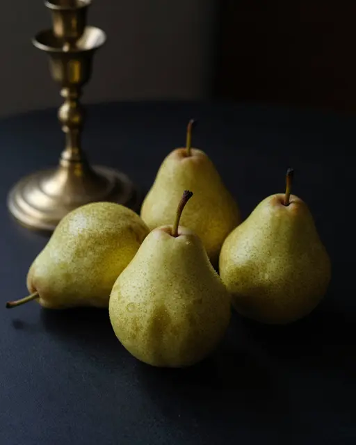 Four fresh pears sit with a brass candlestick on a dark table under low-key lighting