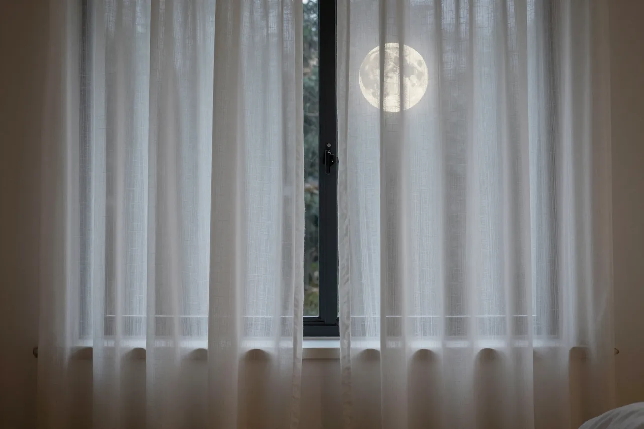 A bright full moon glows behind white sheer curtains covering a bedroom window at night