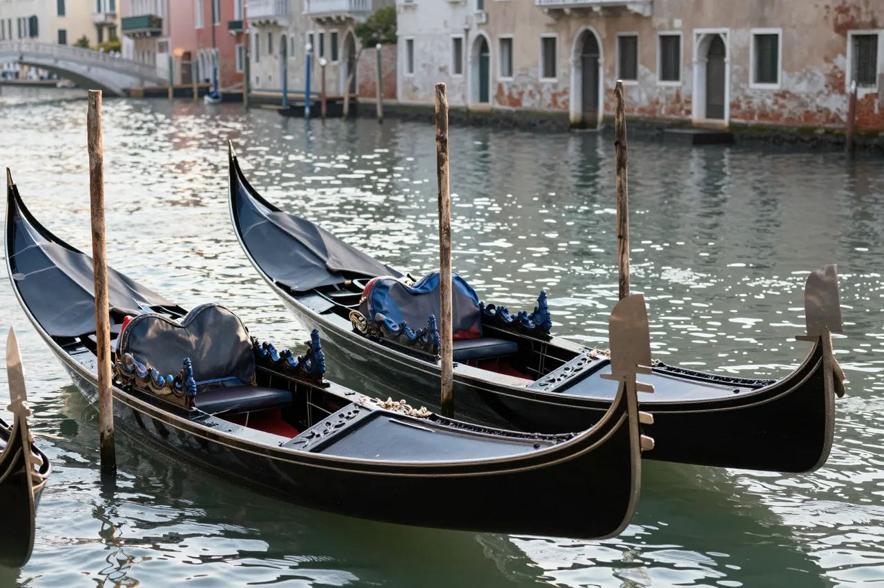 Moored black gondolas rest on sun-dappled Venetian canal water at sunrise, with historic waterfront buildings in the