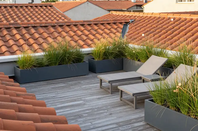 Wooden rooftop terrace with lounge chairs and grass planters overlooking terracotta rooftops at morning