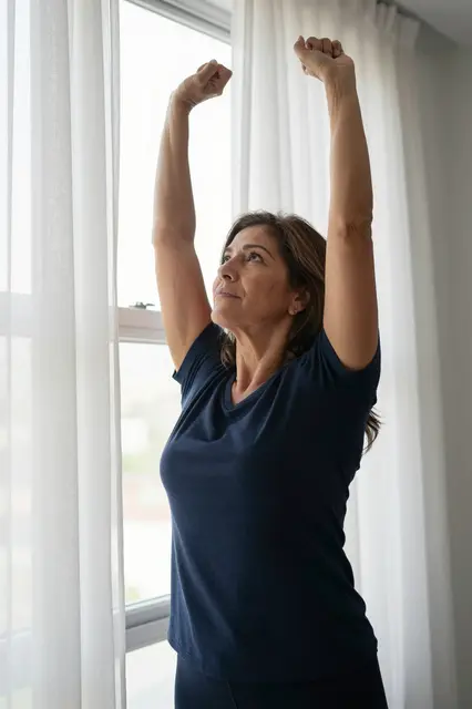 Mid adult woman stretching her arms upward next to a bright window with sheer curtains